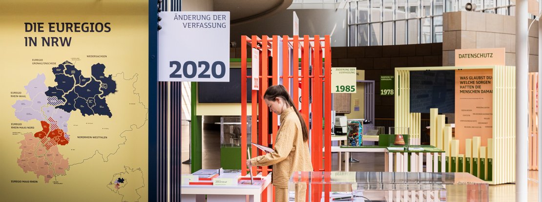 VISUELL Szenografie: Wanderausstellung „Jubiläum. 75 Jahre Verfassung NRW“ für die Stiftung Haus der Geschichte Nordrhein-Westfalen VISUELL Szenografie: Wanderausstellung: Blick in die Ausstellung mit farbigen Modulen und Informationstafeln zu Themen wie Europa, Datenschutz und Verfassungsänderungen. Eine Person betrachtet Exponate zwischen farbigen Rahmen.